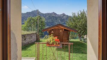 Una baita di legno con parete in pietra e cancello in metallo, immersa nel verde con montagne sullo sfondo.