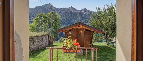 A wooden cabin with a stone wall and a metal gate, surrounded by greenery and mountains in the background.