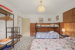 Una habitació amb una llit doble, un armari de fusta, una prestatgeria amb roba, una làmpada penjant i una porta.
