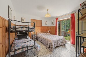Une chambre avec un lit superposé, une armoire en bois, un lit recouvert d’une couvre-lit à motifs floraux, un luminaire suspendu et une vue sur la verdure par la fenêtre.