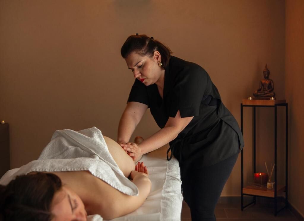 Eine Massageanwendung in einem Raum mit Massageliege, Statue und einer kleinen Ablage mit Kerzen.