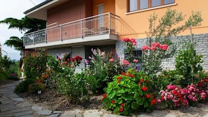 A two-story house with a balcony, surrounded by a garden with various flowering plants. - Casa SOLE, swimming pool and wellness (Castiglione Tinella)