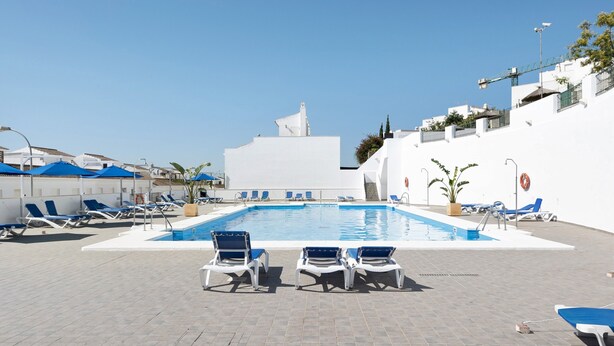 Una piscina con sillas de descanso, cielo despejado y un edificio blanco con una estructura de techo distintiva.