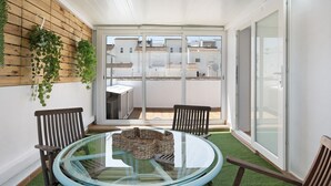 Un balcón con mesa de vidrio, dos sillas, una cesta tejida y una pared de madera con plantas colgantes.
