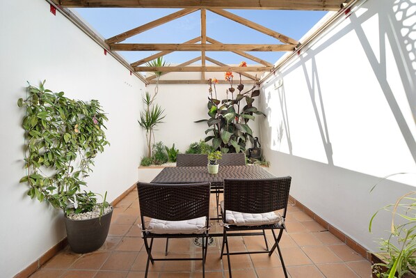 Een overdekt terras met een tafel en stoelen, omringd door potplanten en een houten dakconstructie.