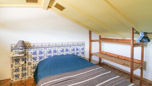 A small, single-bed room with a sloped ceiling, a wooden bunk bed, a bedside lamp, and a tiled wall with a decorative pattern.