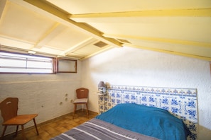 A small, single-room with a sloped ceiling, a bed with a blue headboard, a chair, and a small table with a lamp.