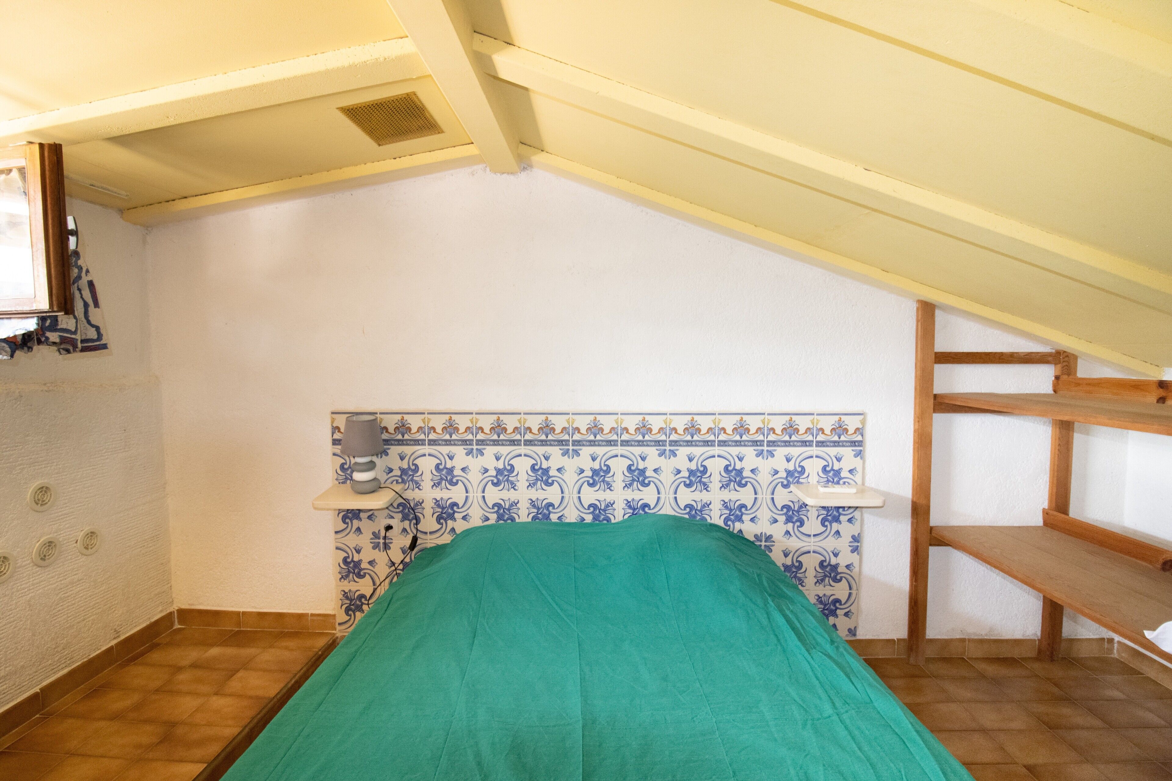 A small, single-bed room with a sloped ceiling, a tiled backsplash, and a wooden shelf.