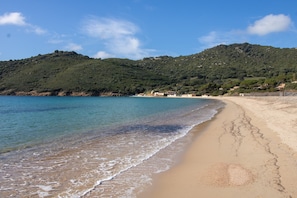Une plage avec une eau claire, un rivage sablonneux et une colline où se trouvent des bâtiments en arrière-plan.