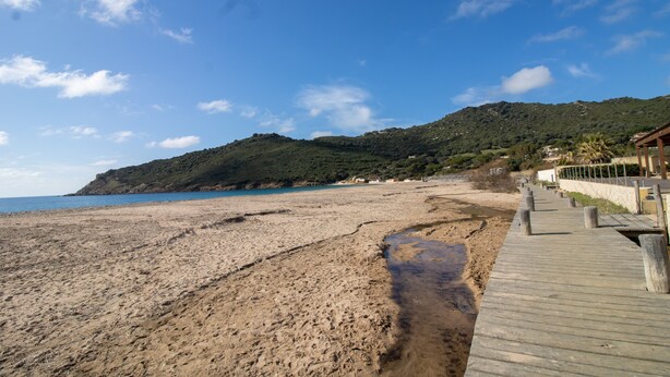 A beach with a wooden boardwalk leading to a building with a view of the sea and hills in the background. - “Le Cinque” Apartment with Sea View, Wi-Fi, and Air Conditioning (Appietto)