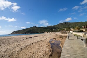 A beach with a wooden boardwalk leading to a building with a view of the sea and hills in the background.