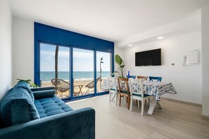 A modern living room with a large window, a blue sofa, a dining table with chairs, and a flat-screen TV mounted on the wall.