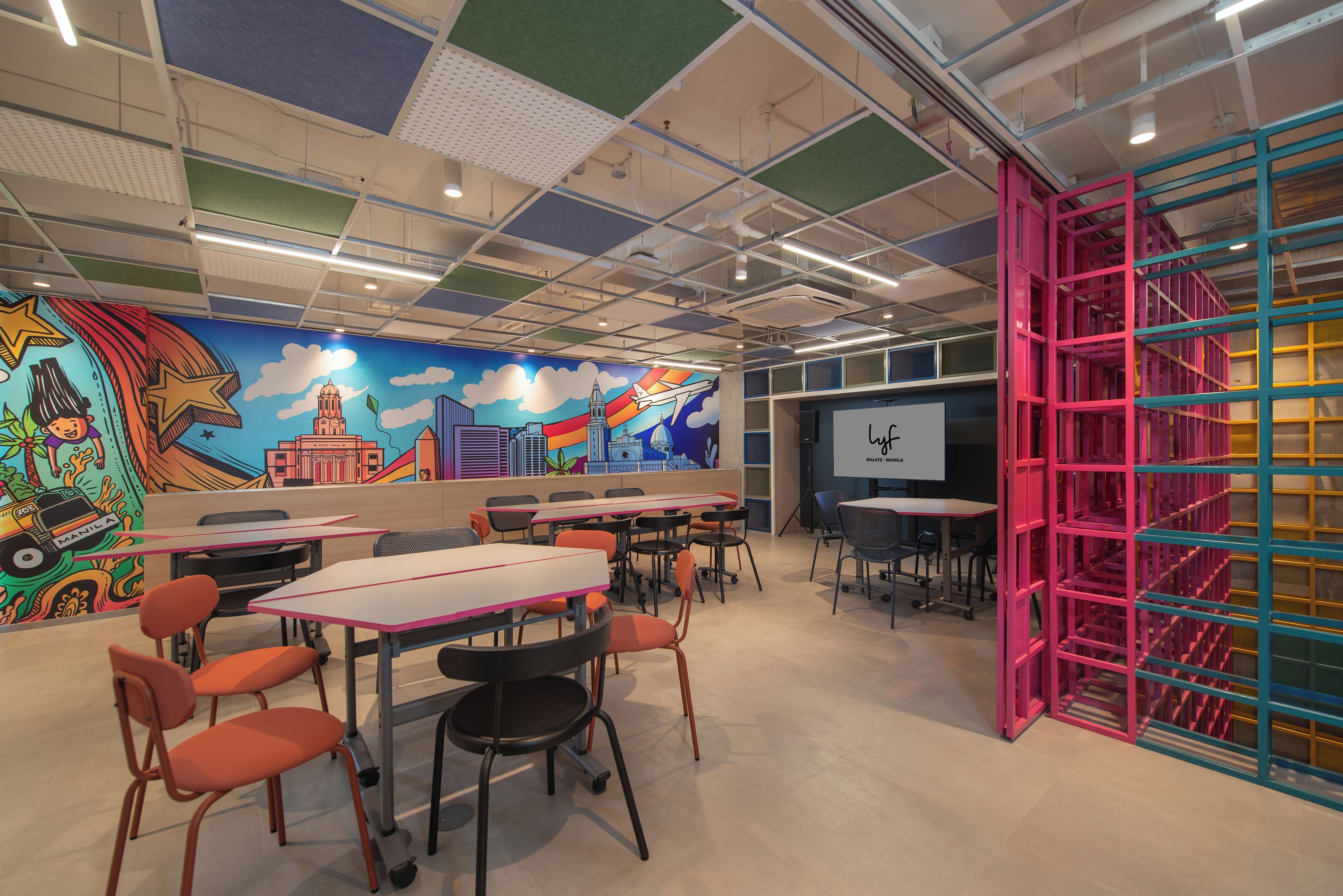 A classroom with tables and chairs, a colorful mural, and a pink and blue partition.