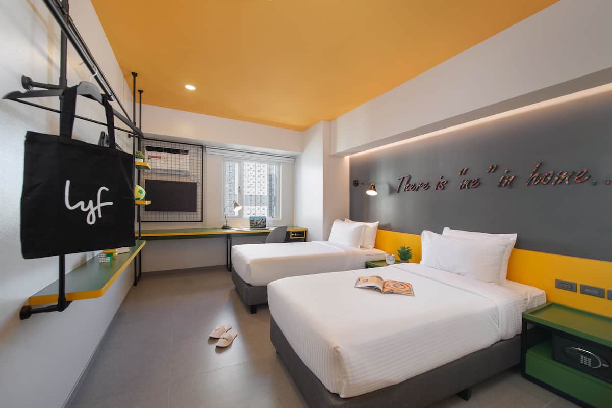 Quarto de hotel moderno com duas camas, uma escrivaninha, uma TV e uma parede com uma frase motivacional.