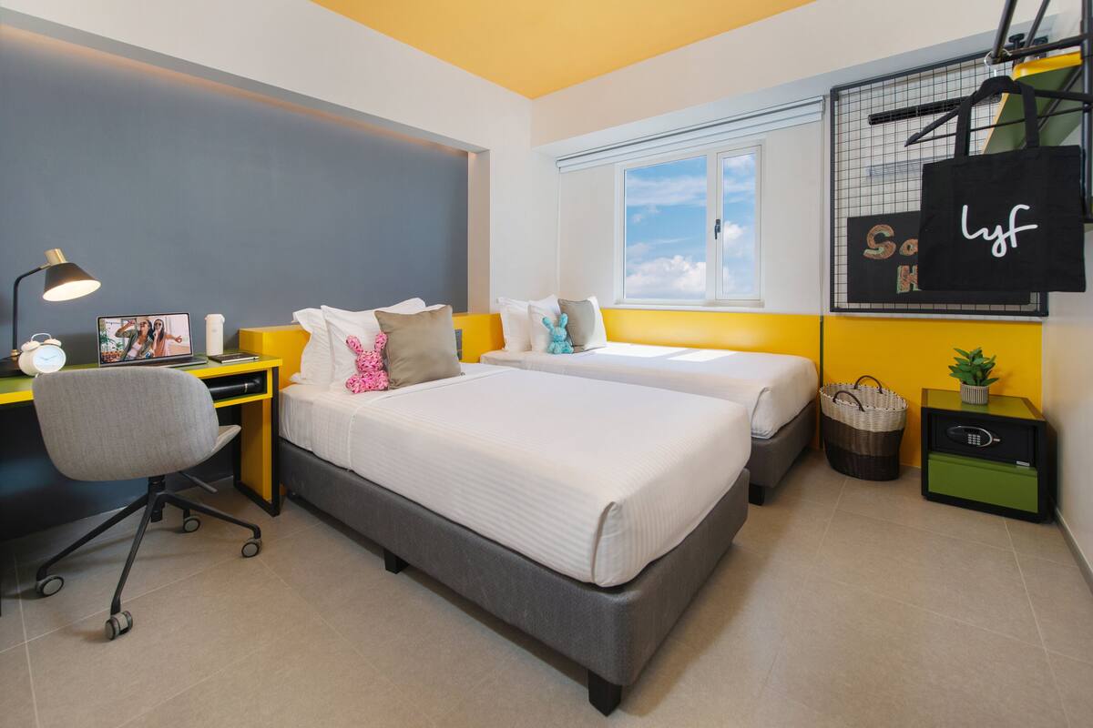 Quarto moderno com duas camas, uma escrivaninha, uma cadeira, uma luminária, um cesto e uma pequena planta.