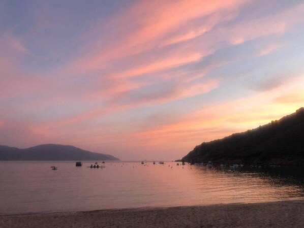 A calm body of water with boats, a beach, and a hilly landscape in the background.