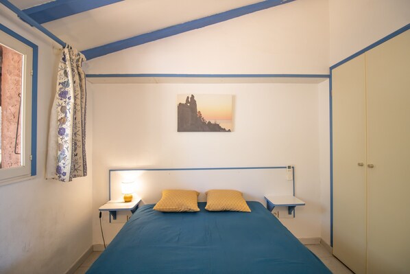 A bedroom with a bed, two bedside tables, a lamp, a window with curtains, a wall-mounted painting, and a wardrobe.
