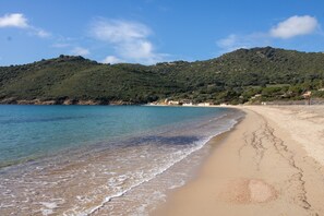 A beach with clear water, a sandy shore, and a hill with buildings in the background.