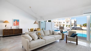 A modern living room with a sofa, armchair, and a view of a pool through large windows. - Vacation Home "Vilamoura Com Piscina E Wifi" with Private Pool, Terraces & Wi-Fi (Vilamoura)