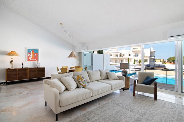 Un salon moderne avec un canapé, un fauteuil et une vue sur une piscine, visible à travers de grandes fenêtres.