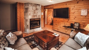 A cozy cabin interior with a stone fireplace, wooden walls, a flat-screen TV, and a comfortable seating area.