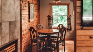 A wooden interior with a round table and four chairs, a window with plaid curtains, and a hanging light fixture.