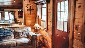 A cozy cabin interior with a wooden interior, a sofa with a blanket, a lamp, and a window with curtains. - Romantic Cabin | Private Stay | King Bed | Fire Pit (Rockbridge)