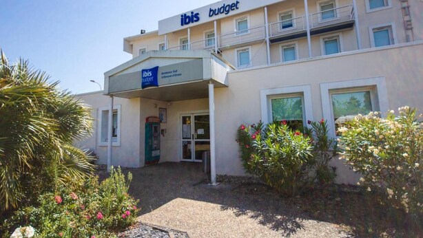 A hotel named 'ibis budget' with a signboard, a covered entrance, and a small garden area with plants and flowers.