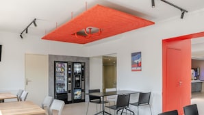 A modern interior with a meeting table, chairs, a vending machine, and a red accent wall. - ibis budget Bordeaux Sud Villenave-d'Ornon (Villenave-d'Ornon)