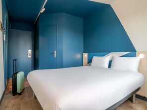 A hotel room with a large bed, a door, a suitcase, and a light fixture. - ibis budget Bordeaux Sud Villenave-d'Ornon (Villenave-d'Ornon)