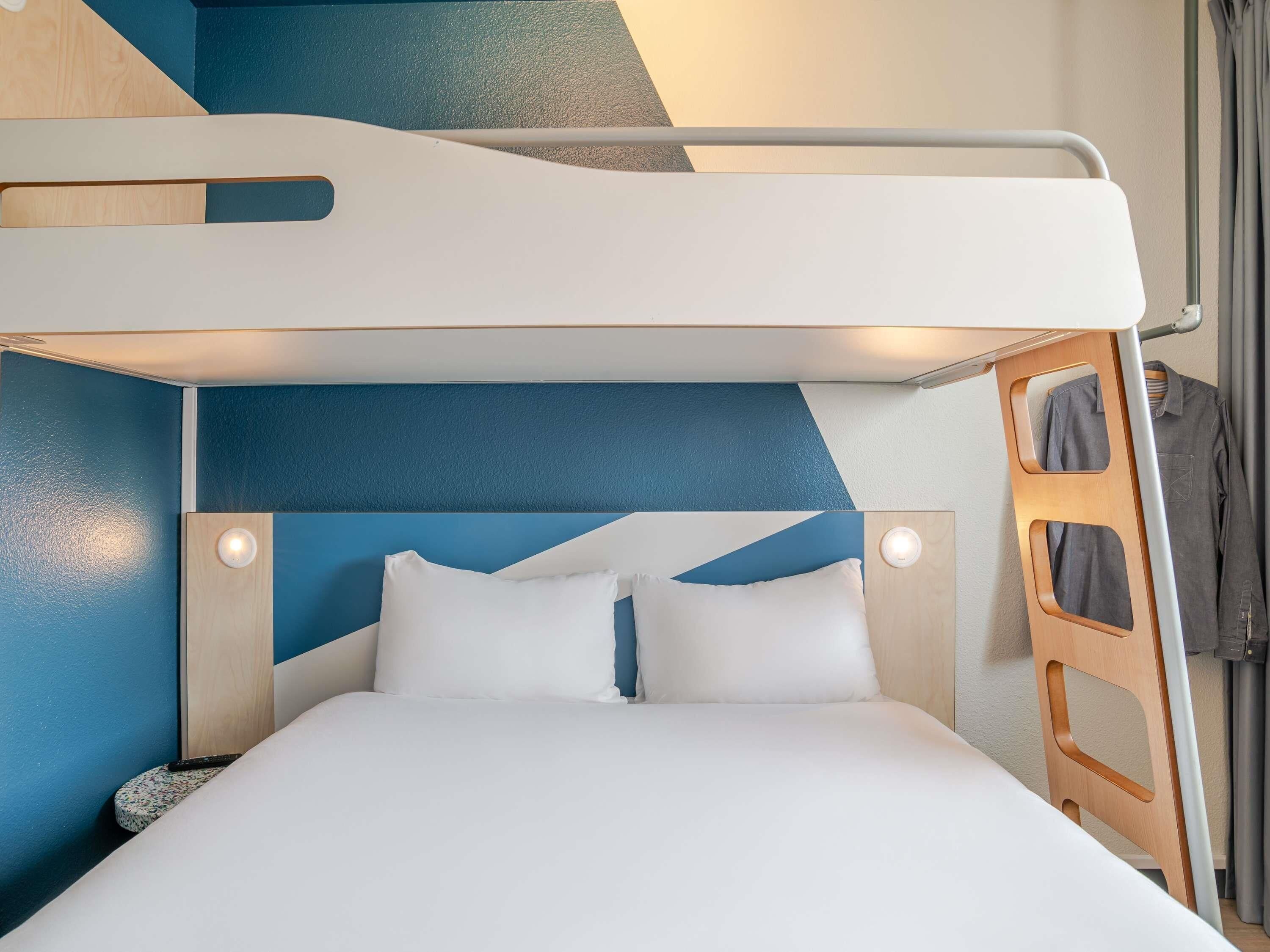 A bunk bed with a white mattress and two pillows, a wooden headboard, and a small bedside table with a lamp.