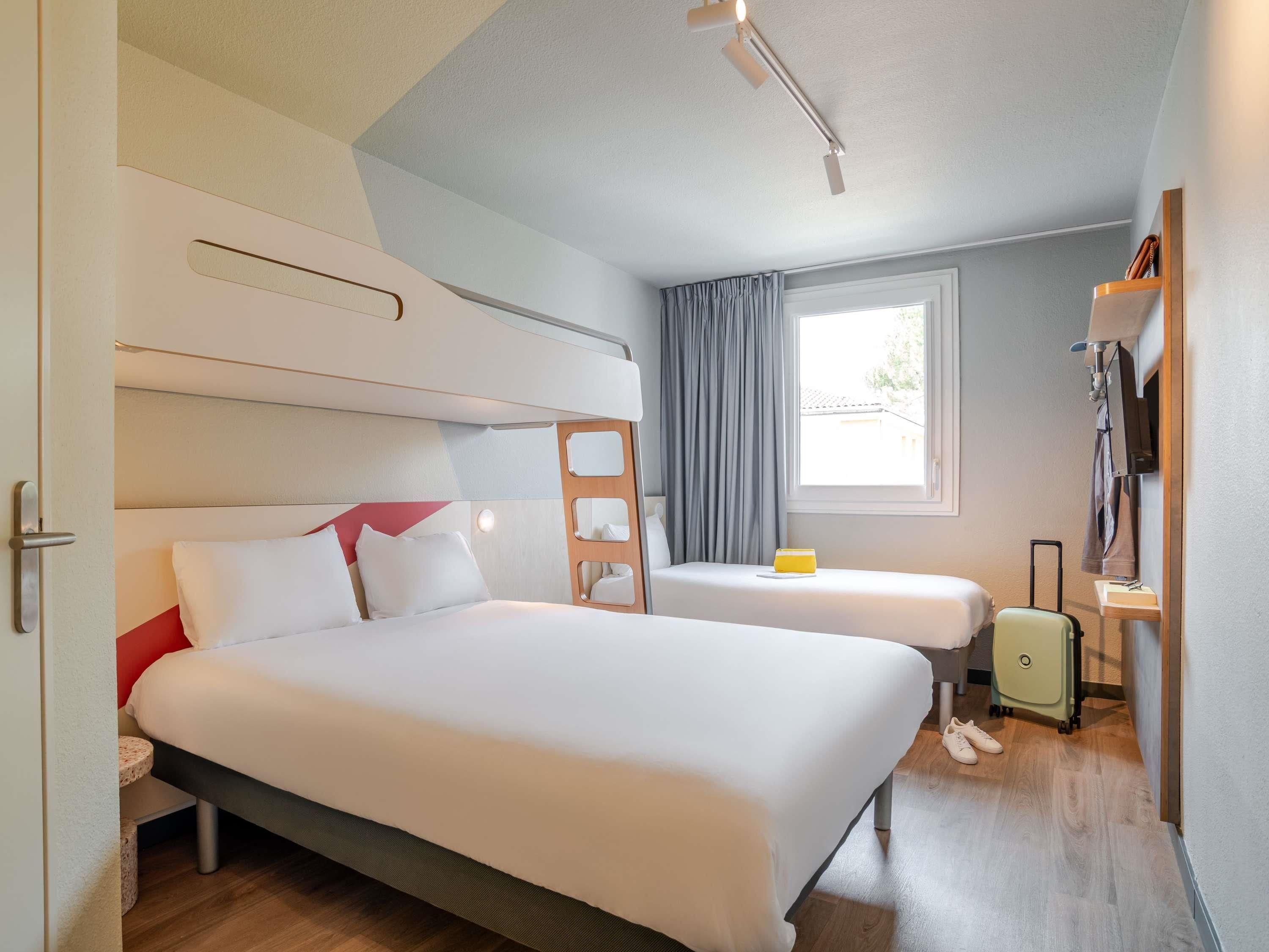 Una habitación de hotel con una cama, una mesita de noche, una ventana con cortinas, una maleta y un pequeño escritorio con una lámpara.