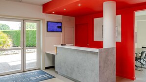 A reception area with a red accent wall, a reception desk, a computer monitor, and a door with a window. - ibis budget Bordeaux Sud Villenave-d'Ornon (Villenave-d'Ornon)
