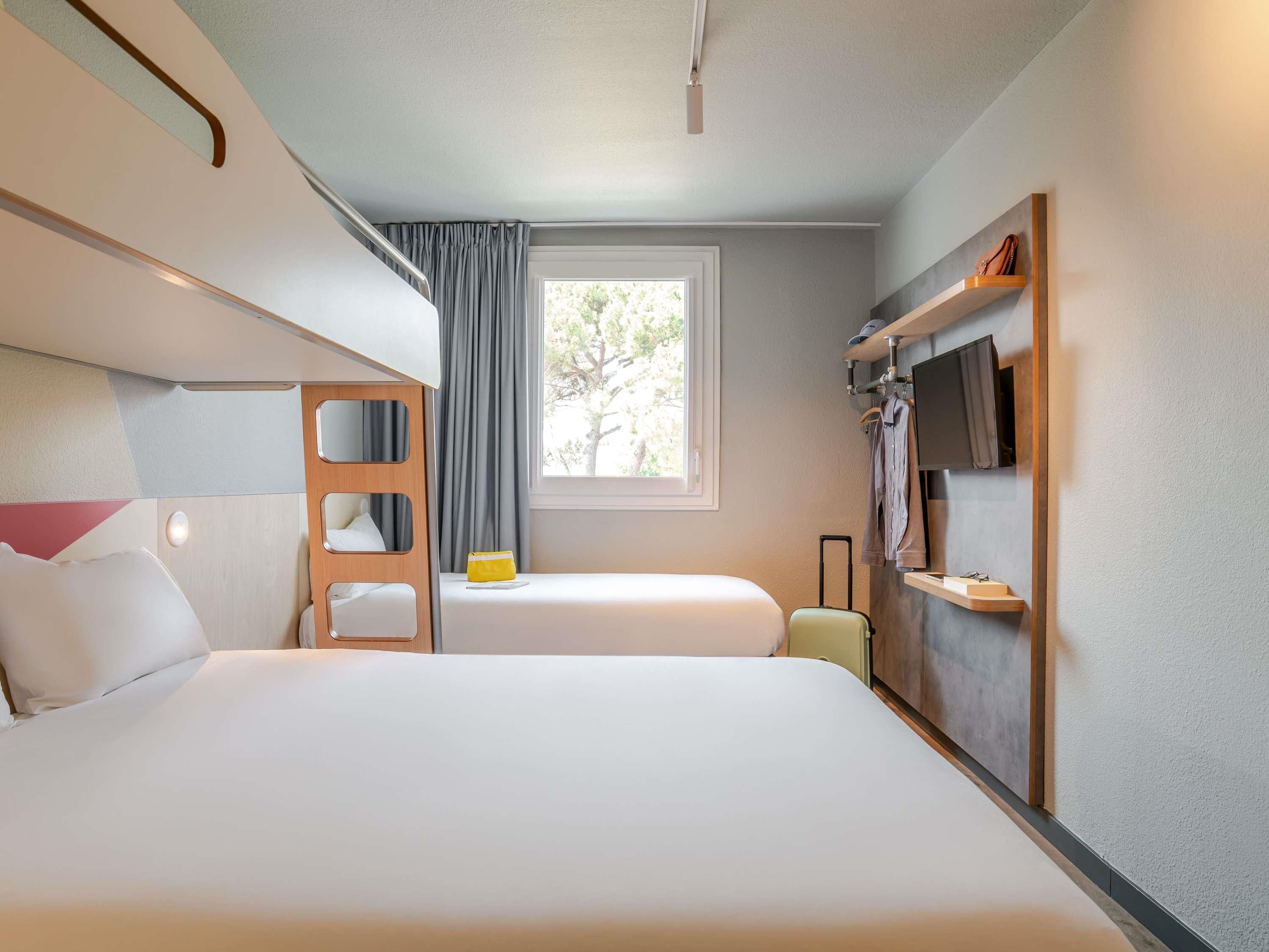 Een hotelkamer met een stapelbed, een enkel bed, een raam met gordijnen, een televisie en een klein tafeltje met een koffer.