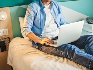 Eine Person sitzt auf einem Bett mit einem Laptop, einem Kopfkissen und einem Nachttisch mit einer Lampe.