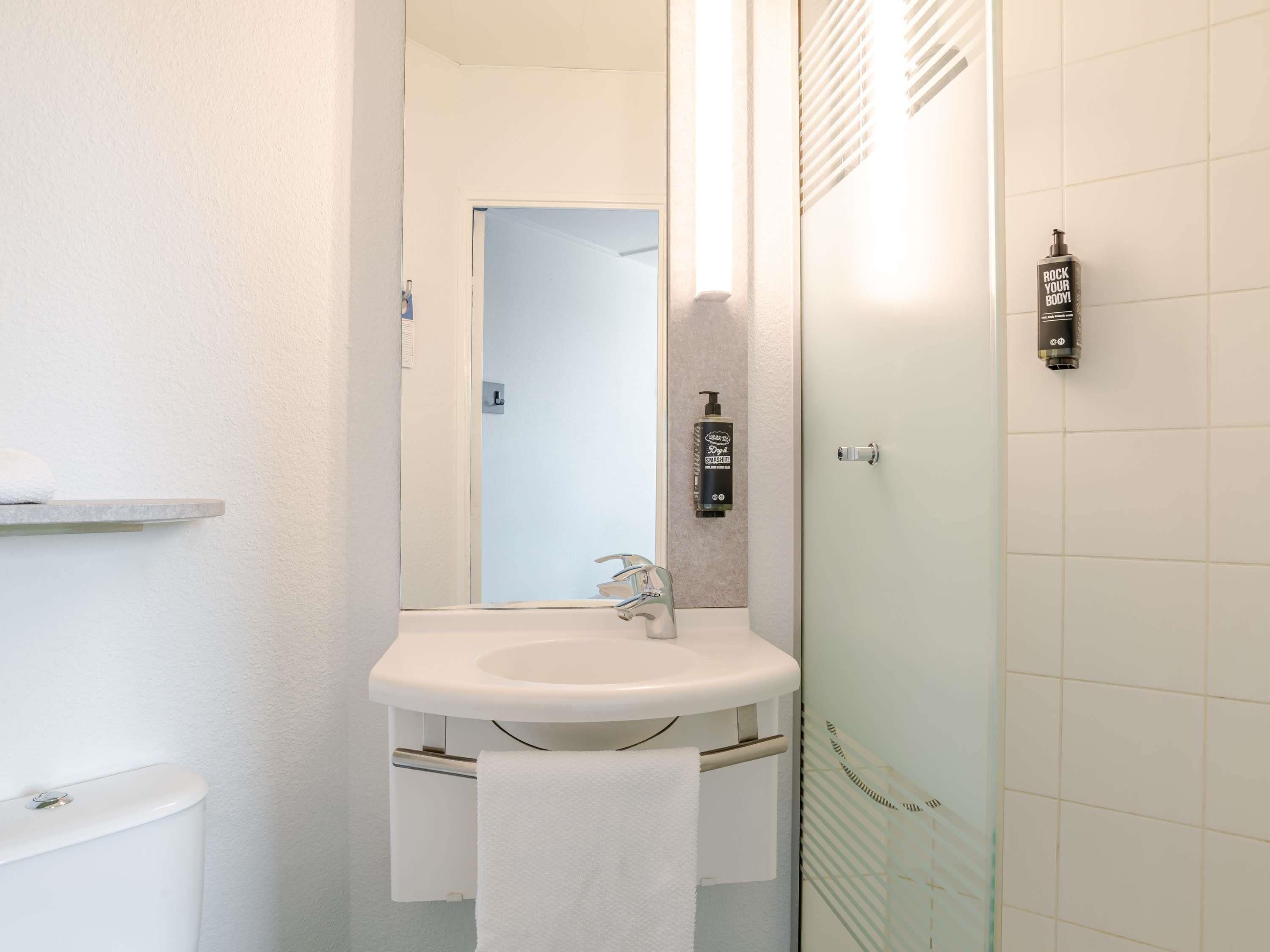 Un baño con lavamanos blanco, espejo, toalla y zona de ducha.