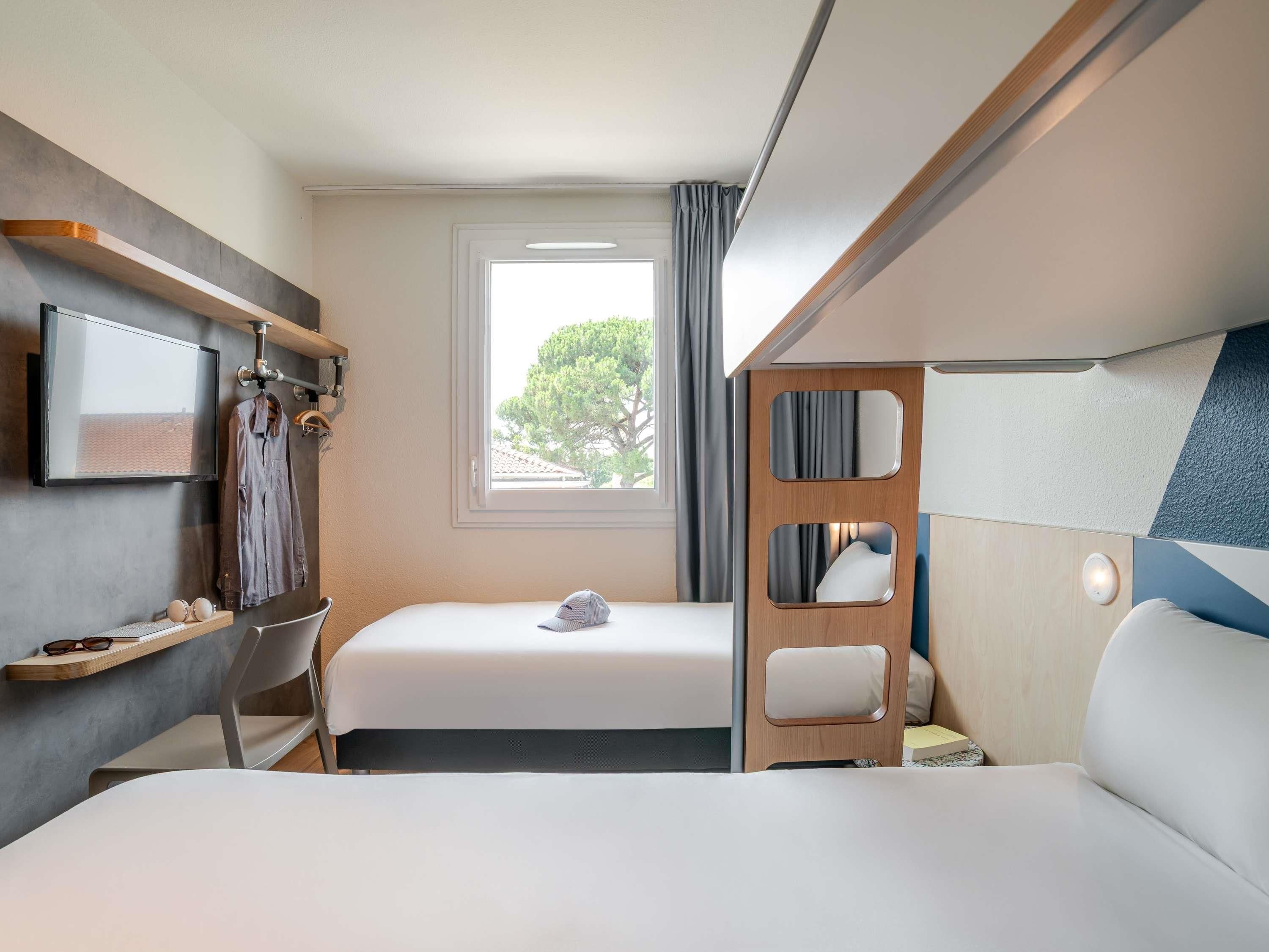 A compact hotel room with a bed, a desk, a chair, a TV, and a window with a view of trees.