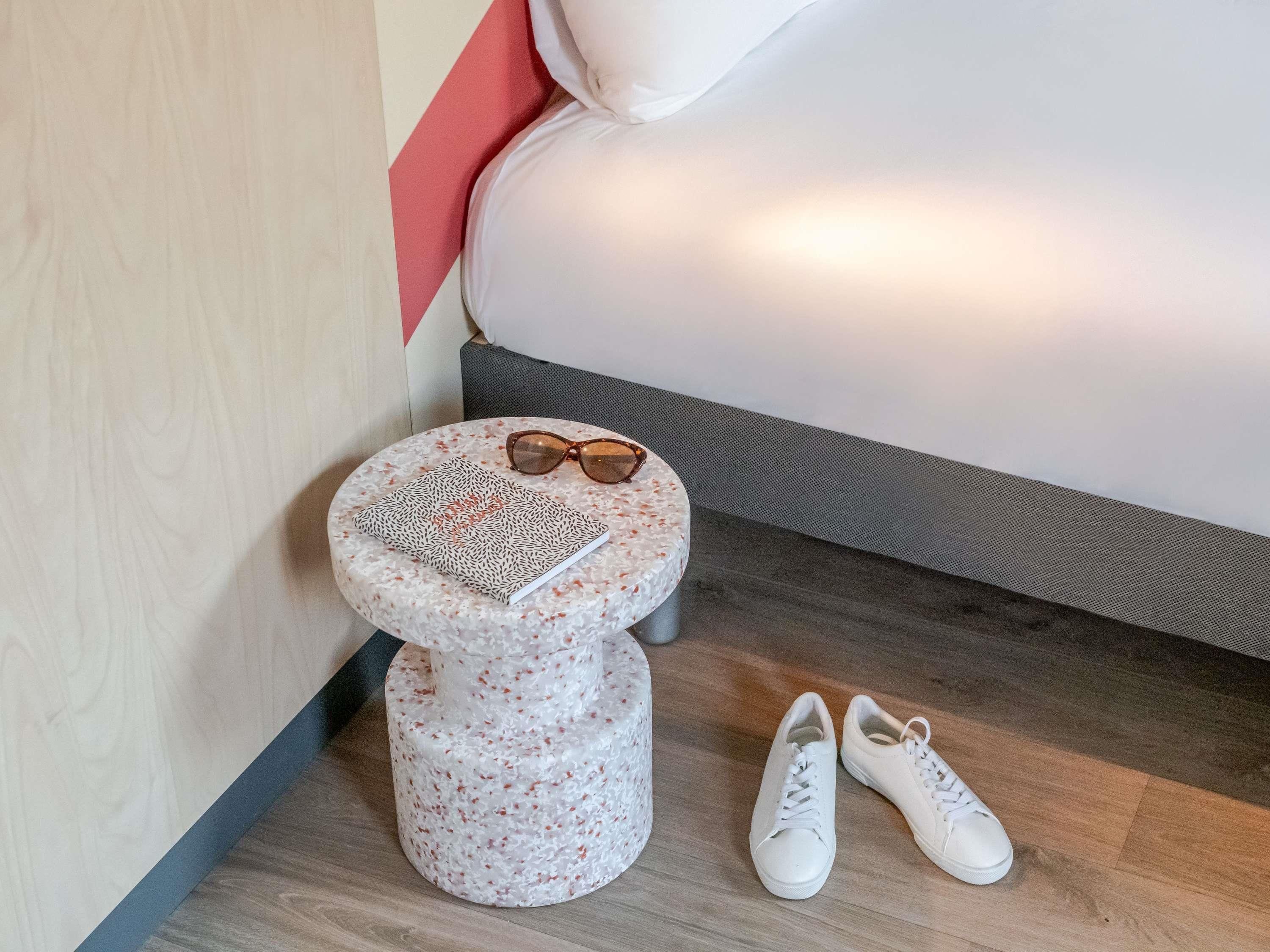 A small round table with a notebook and sunglasses, next to a bed with a grey headboard.
