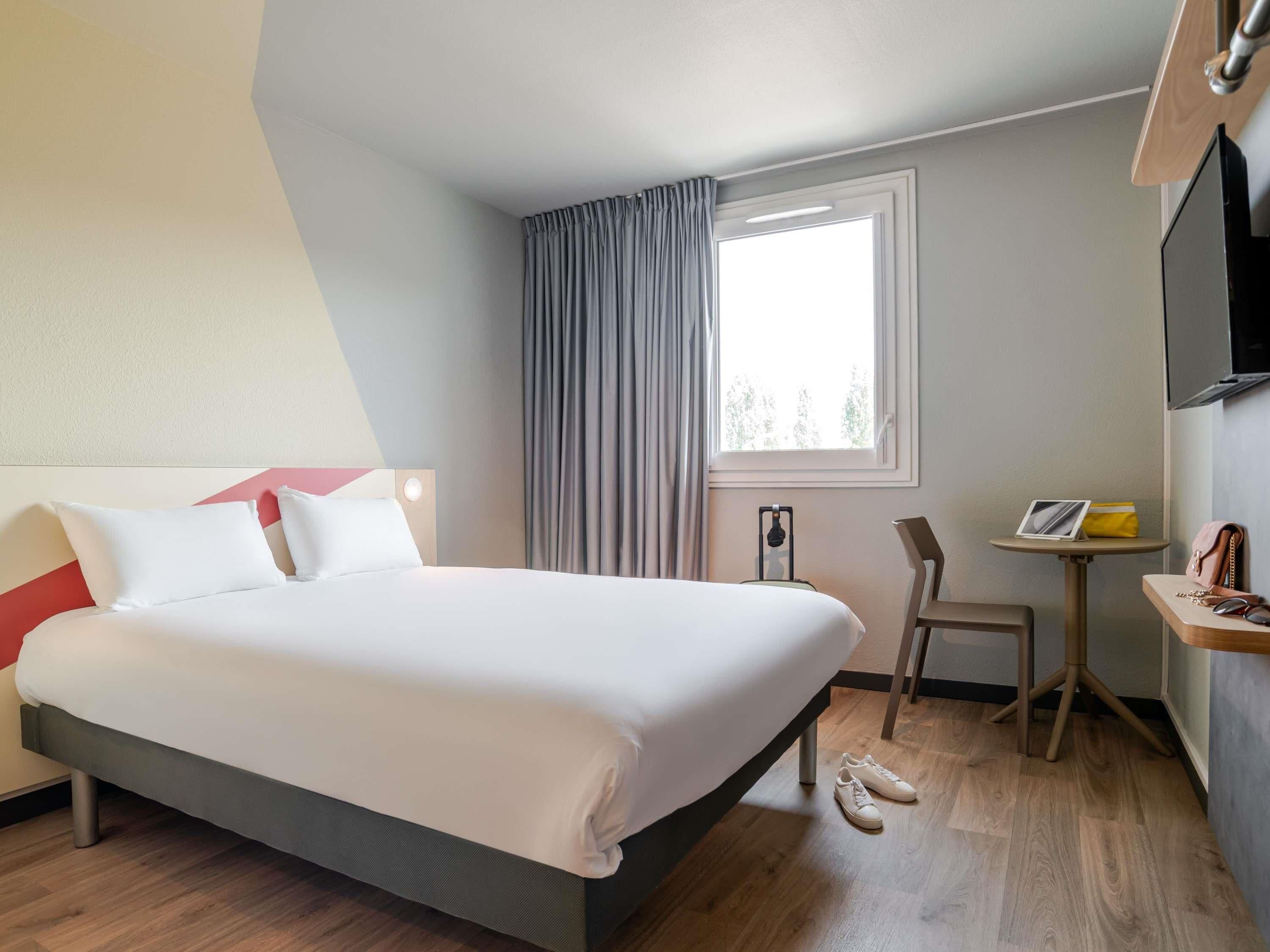 A hotel room with a bed, a chair, a small table, a window with curtains, and a TV mounted on the wall.