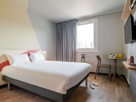 Habitación de hotel con una cama, una silla, una mesita, una ventana con cortinas y un televisor montado en la pared.