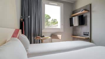 Een hotelkamer met een bed, een klein tafeltje met een boek en een handtas, een stoel, een aan de muur bevestigde televisie en een raam met gordijnen.