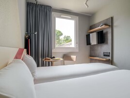Habitación de hotel con cama, una mesita con un libro y un bolso, una silla, televisor montado en la pared y una ventana con cortinas.