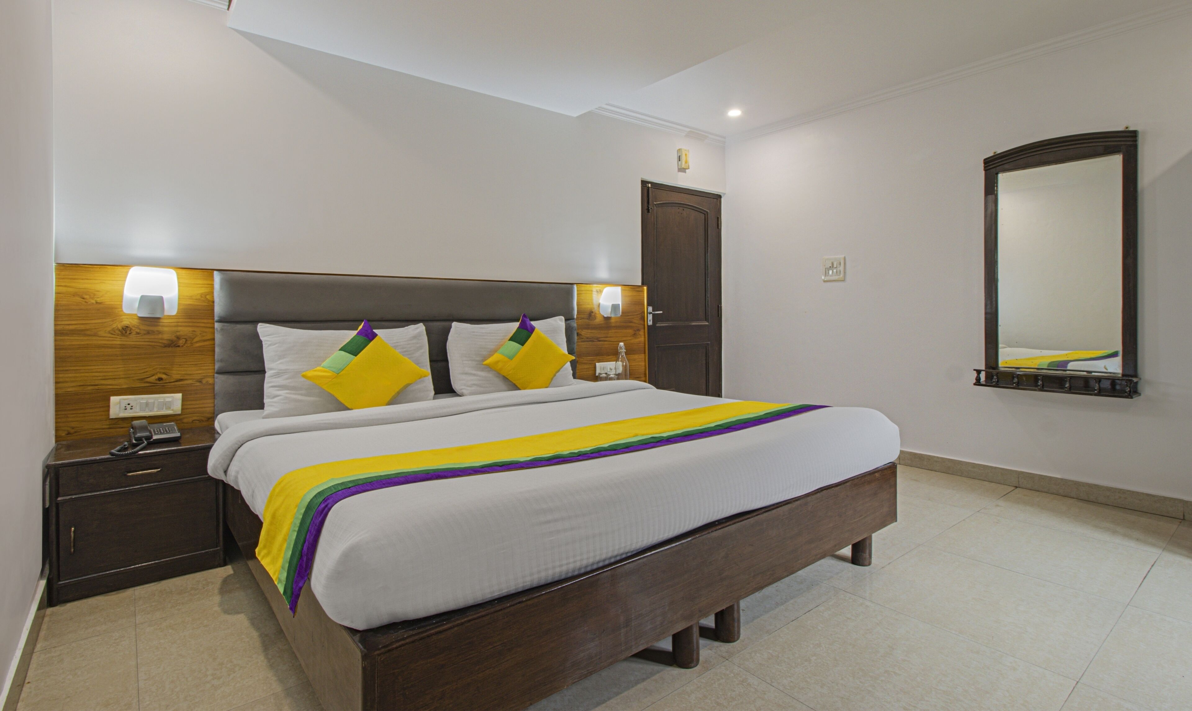 A neatly made bed with a white and colorful bedspread, two bedside tables with lamps, a wooden headboard, a door, and a large mirror on the wall.