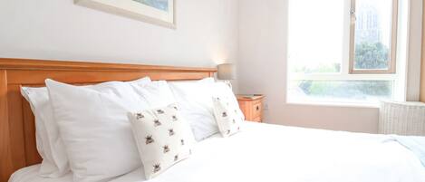 A neatly made bed with white linens and patterned pillows, a wooden headboard, a bedside table with a lamp, and a window with a view of greenery.