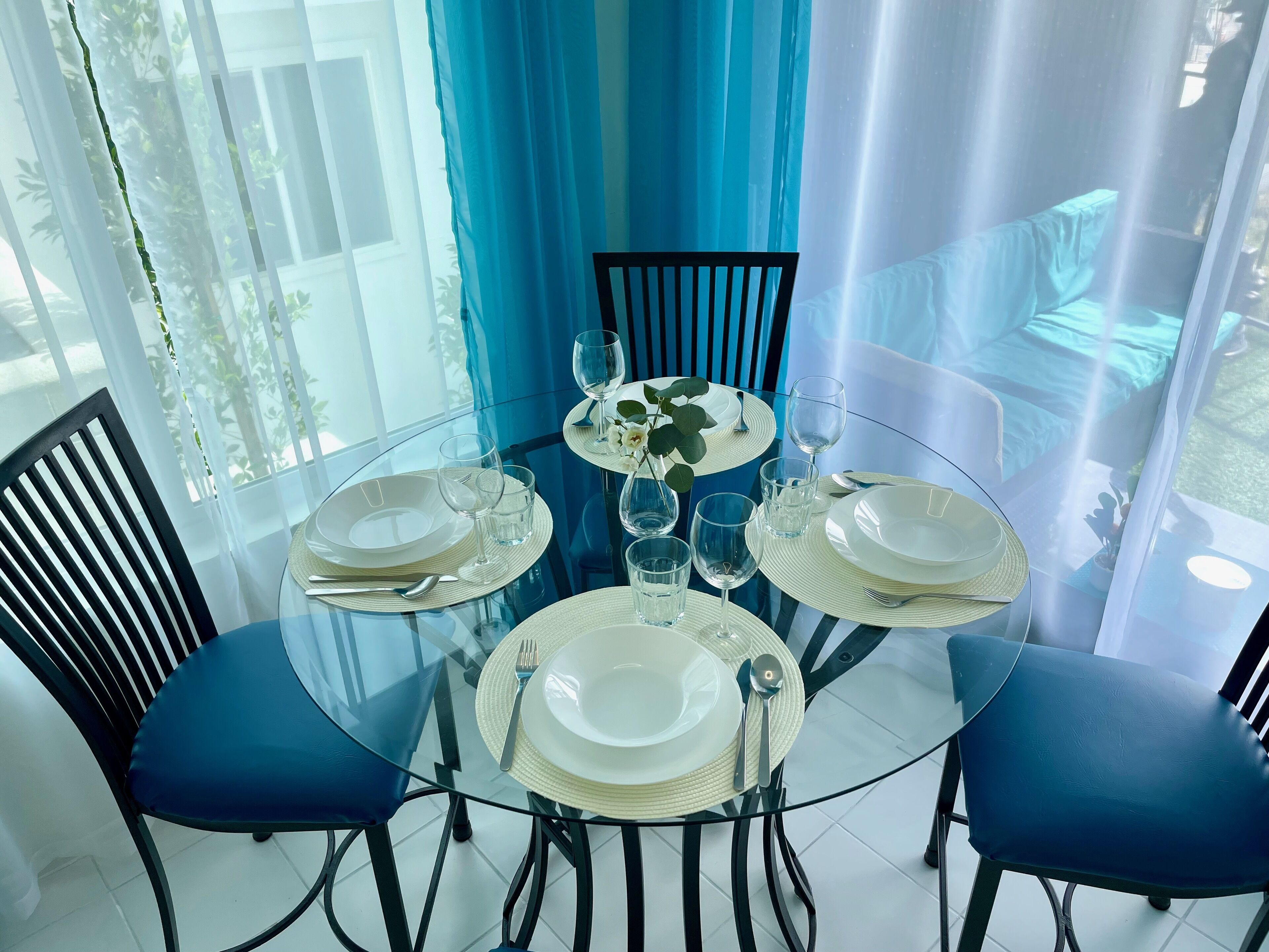A dining table set for four with clear glassware and plates, surrounded by chairs with blue cushions.