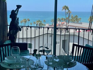 A dining table with plates, glasses, and cutlery, overlooking a beach with palm trees and a statue. - SurfView VR: Best Ocean and Pier View, 5 mins to Beach, Cozy Patio, Pet Friendly (San Clemente)