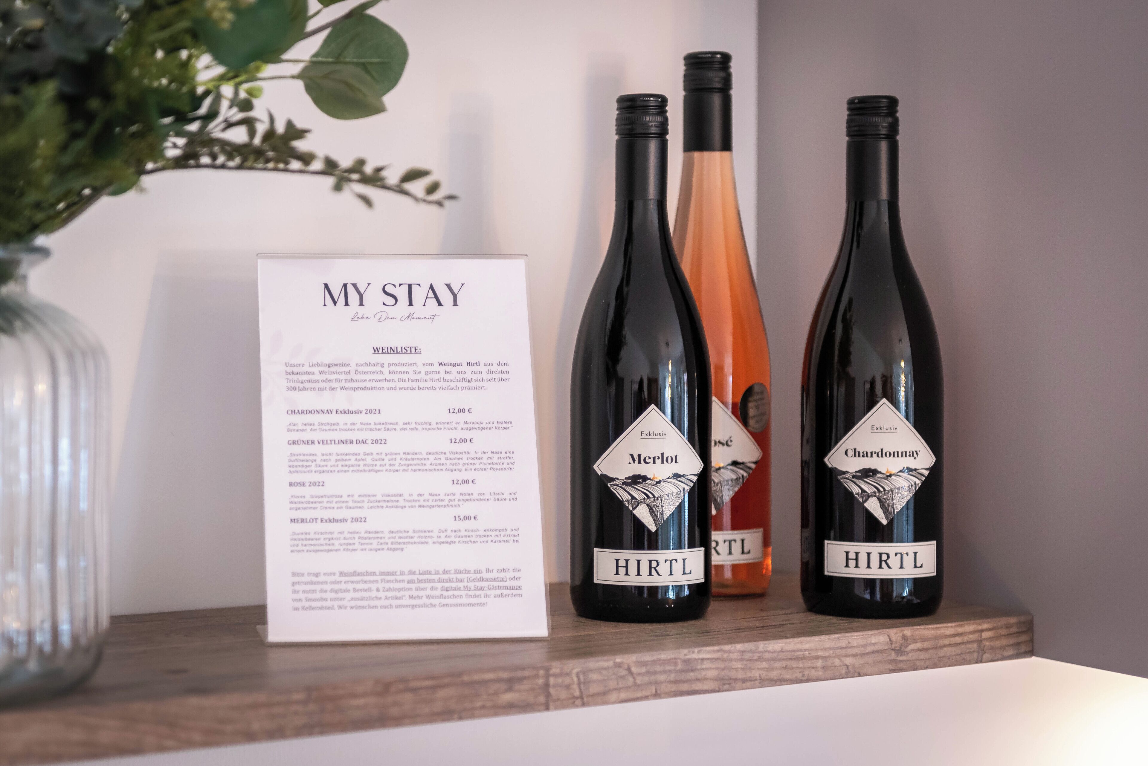 Twee flessen wijn, gelabeld 'HIRTL' en 'Merlot', op een houten plank met een menukaart met de titel 'MY STAY'.
