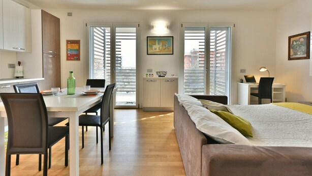 A compact hotel room with a bed, a desk, a chair, and a dining area. - GuestHost - Casa Kikka: luminoso bilo con balcone (Cernusco sul Naviglio)