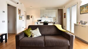 A modern living room with a sofa, a dining area, and a kitchen in the background. - GuestHost - Casa Kikka: luminoso bilo con balcone (Cernusco sul Naviglio)