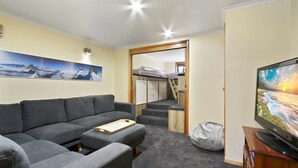 Un espace de vie compact comprenant un canapé, un téléviseur, un lit superposé et un escalier menant à un coin cuisine.
